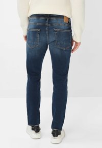 Uomo in piedi con le mani ai fianchi, indossando jeans blu scuro, un maglione bianco lavorato a maglia e sneakers bianche con dettagli neri nel tallone.