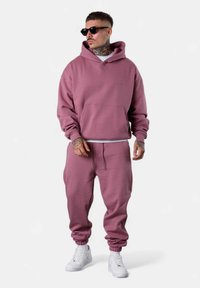 Pink hoodie and matching sweatpants made of soft fabric. The hoodie features a front pocket, elastic cuffs, and a relaxed fit. White sneakers.