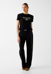 Frau steht, trägt ein schwarzes GUESS-T-Shirt und schwarze Hosen mit beigen Paspeln, kombiniert mit braunen Schuhen, schaut leicht zur Seite.