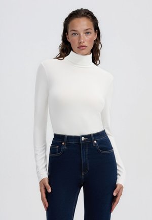 Donna con capelli mossi indossa un dolcevita bianco aderente e jeans blu scuro a vita alta su uno sfondo chiaro e uniforme.