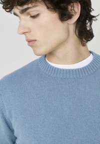 Pull en tricot bleu clair avec un col côtelé et une texture, présentant un design ajusté. L'arrière-plan est blanc uni.