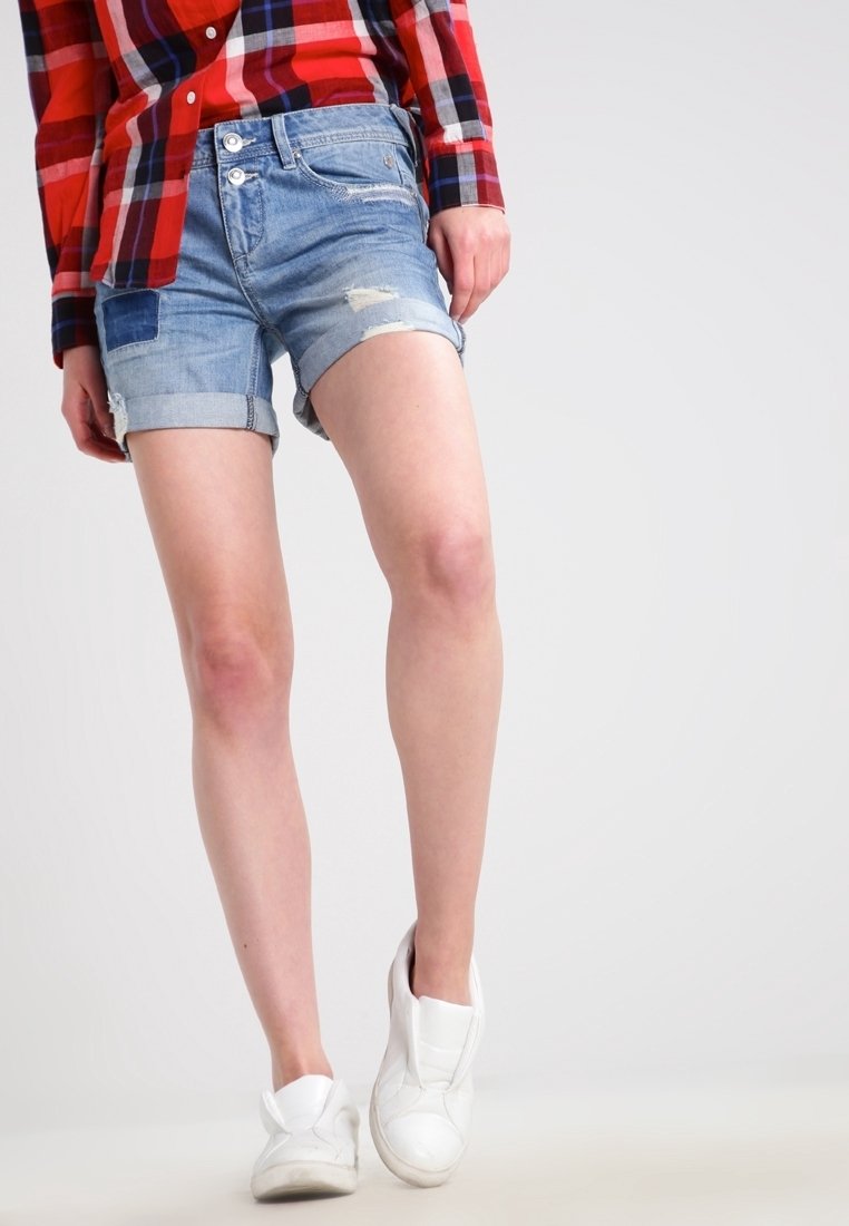 Jasnoniebieskie szorty dżinsowe z postarzanymi detalami, podwiniętym dołem i metalowymi guzikami, zestawione z białymi trampkami i czerwoną koszulą w kratę.