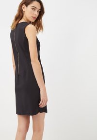 Mujer con un vestido negro sin mangas y una cremallera dorada visible en la espalda, de pie y mirando por encima del hombro.