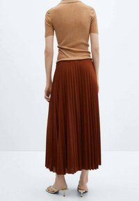 Persona vestida con una blusa corta de manga corta color tostado con textura acanalada, una falda midi plisada color óxido y sandalias de tacón de kitten beige, de pie sobre un fondo liso.