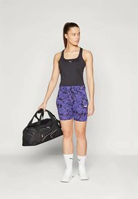 Kvinne som står i treningstøy og holder en svart treningsbag, iført svart tank topp, lilla mønstrede shorts, hvite sokker og hvite joggesko.