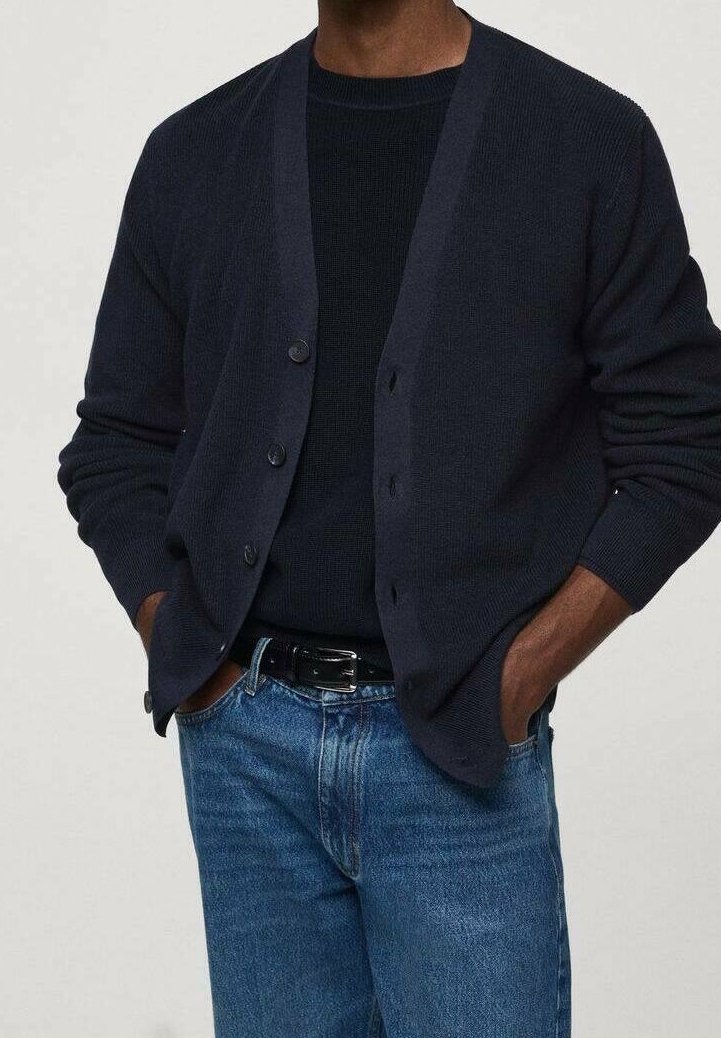 Homme portant un cardigan bleu marine foncé boutonné, un pull bleu marine, un jean bleu et une ceinture noire, debout les mains dans les poches.