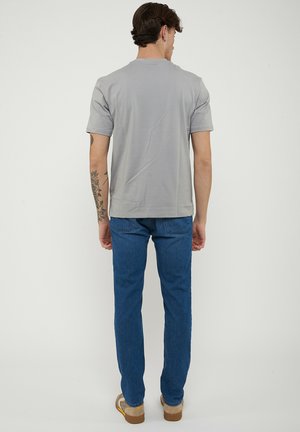 Uomo con braccio tatuato che indossa una maglietta grigia a maniche corte, jeans blu e sneaker beige, in piedi con la schiena rivolta verso uno sfondo semplice.