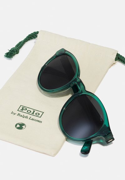 Óculos de sol verdes, transparentes, com lentes oversized, repousando sobre uma bolsa de tom bege claro com texto verde e um logótipo, com fecho de cordão.