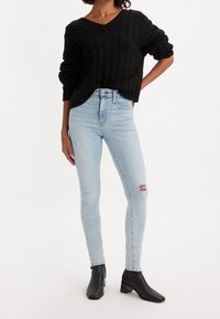 Suéter negro de punto con un patrón de trenzas, jeans ajustados rasgados de color azul claro y botas negras hasta el tobillo con punta cuadrada y tacón ancho.