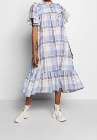 Osoba oblečená v volných, řasených modro-bílých kostkovaných midi šatech s nabíranými rukávy a bílými teniskami, stojící na jednorázovém šedém pozadí.
