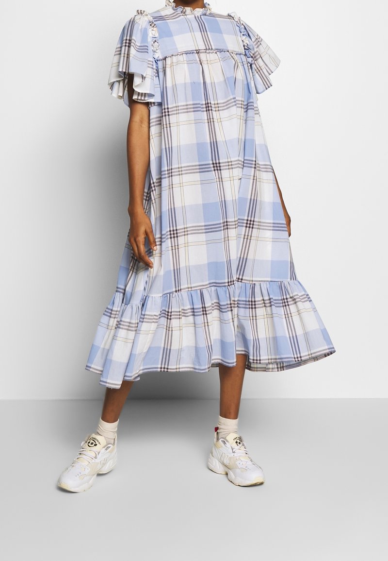 Osoba oblečená v volných, řasených modro-bílých kostkovaných midi šatech s nabíranými rukávy a bílými teniskami, stojící na jednorázovém šedém pozadí.