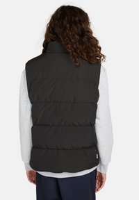 Schwarze gepolsterte Weste mit hohem Kragen, die über eine gesteppte Textur und eine glatte Oberfläche verfügt. Getragen über einem grauen Langarmshirt.