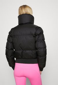 Schwarze Daunenjacke mit hohem Kragen, gestepptem Muster und geripptem Saum. Getragen über leuchtend pinken Leggings.