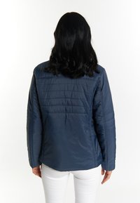Giacca trapuntata blu navy con finitura liscia, caratterizzata da una chiusura a zip frontale, collo rialzato e cuciture invisibili. Pantaloni bianchi visibili sotto.