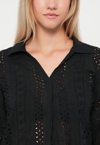 Black blouse featuring a collared neckline, v-neck design, and intricate eyelet embroidery with floral patterns. Soft texture and breathable material.