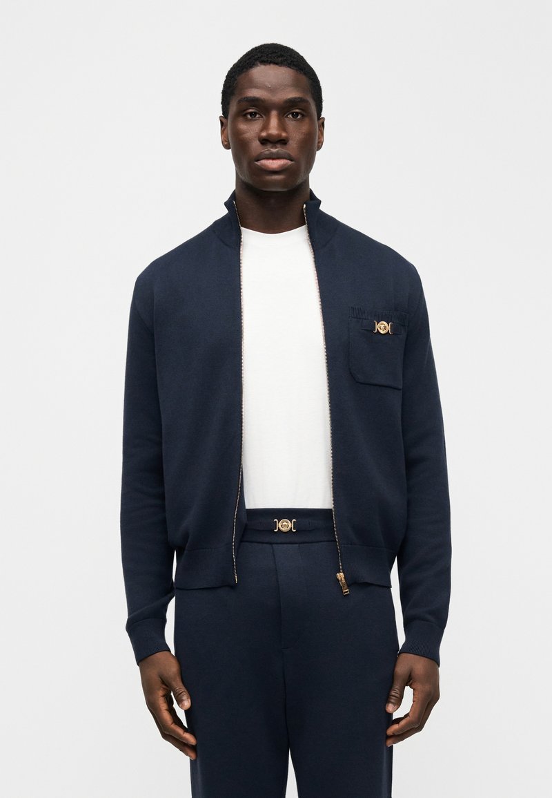 Jeune homme portant un cardigan à fermeture éclair bleu marine sur une chemise blanche, un pantalon bleu marine assorti, debout devant un fond clair uni.