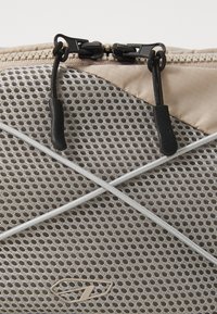 Mesh storage bag in beige with a textured finish. Gray accent lines cross the front. Features dual black zipper pulls for closure.