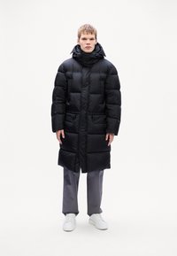 Sort puffjakke med et langt design, store quiltede sektioner, høj krave og forlommer. Sammen med grå bukser og hvide sneakers.