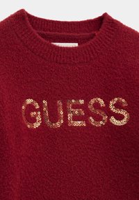 Burgundijas džemperis ar teksturētu virsmu un apaļu dekoltē, ar zeltītām sekvencēm, kas uzrakstītas "GUESS" krūšu daļā.