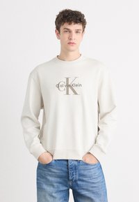 Svijetlosiva dukserica s dugim rukavima koja ima veliki smeđi "CK" logo s natpisom "Calvin Klein" ispod. Mekana tekstura s rebrastim manžetama i donjim rubom.