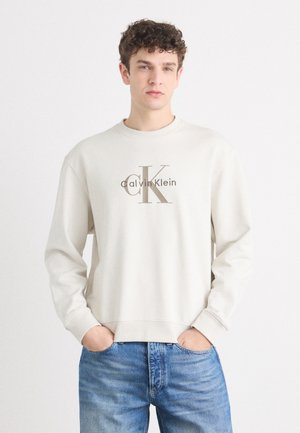 Svijetlosiva dukserica s dugim rukavima koja ima veliki smeđi "CK" logo s natpisom "Calvin Klein" ispod. Mekana tekstura s rebrastim manžetama i donjim rubom.