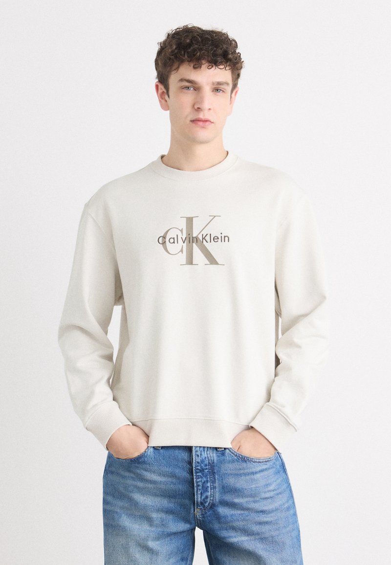 Svijetlosiva dukserica s dugim rukavima koja ima veliki smeđi "CK" logo s natpisom "Calvin Klein" ispod. Mekana tekstura s rebrastim manžetama i donjim rubom.
