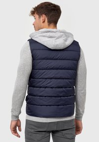 Marineblå tæppevest, quiltet design, polstret med en grå jerseyhætte. Har en glat tekstur og detalje med sideetiket.