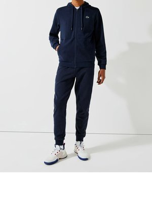 Persoon draagt een marineblauwe zip-up hoodie en bijpassende joggingbroek met witte sportschoenen tegen een effen achtergrond.