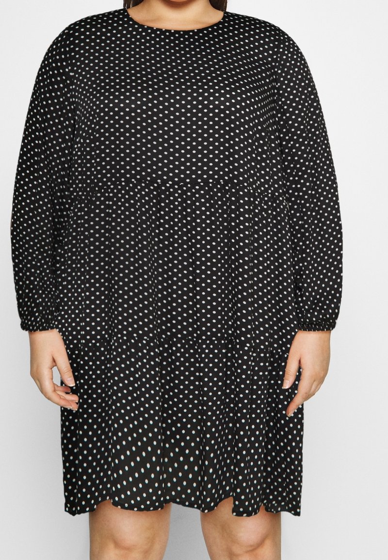 Femme portant une robe noire à manches longues avec des pois blancs, debout, les bras détendus le long du corps.