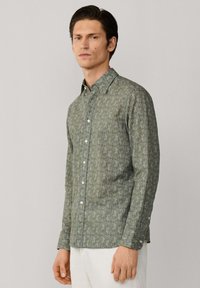 Muž v zelené košili s dlouhým rukávem a bílých kalhotách stojí před jednobarevným světlým pozadím.