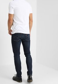 Camiseta blanca de manga corta con cuello redondo combinada con jeans ajustados azul oscuro que tienen bolsillos traseros y un zapato negro resistente.