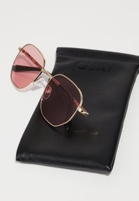 Gafas de sol de gran tamaño con tinte rosa y montura de metal dorado, con almohadillas nasales transparentes, reposando sobre un estuche de piel negra con la palabra "QUAY" grabada.
