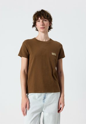 Giovane con capelli corti e ricci che indossa una t-shirt marrone a maniche corte e jeans azzurri su uno sfondo chiaro uniforme.