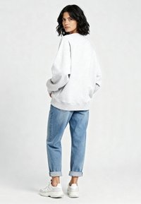 Felpa grigia con vestibilità ampia e polsini a coste, abbinata a jeans azzurri chiari risvoltati e sneakers bianche. Sfondo semplice e neutro.