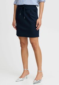 Fustă dintr-un material elastic de culoare bleumarin, cu talie ajustabilă prin șnur, buzunare laterale și un design ajustat, asortată cu pantofi argintii cu vârf ascuțit.