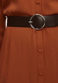 Brun läderbälte med en rund silverspänne, bärs över en orange knappklänning i mjuk tyg. Har sömnadsdetaljer.