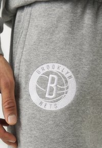 Šedé bavlnené tepláky s okrúhlym bielym vyšívaným logom s textom "BROOKLYN" a "NETS" a so stredným symbolom 'B'.