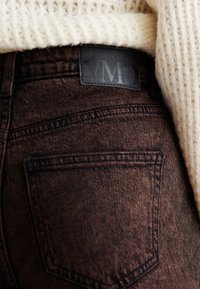 Jeans de ganga castanhos com um acabamento texturizado; apresenta um rótulo de couro preto marcado "VMD" e um design de bolso traseiro. Suéter de malha branca em cima.