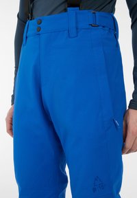 Blauwe skibroek van duurzame stof, met twee knopen, verstelbare bretels en ritszakdetails. Geweven logo nabij de zoom.