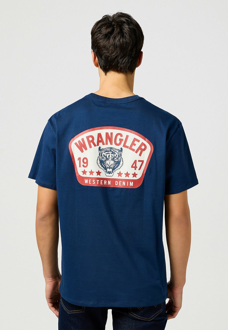 Tummansininen puuvillapaita, jossa on suuri printtipaikka selässä, jossa on tiikeri-kuvio ja teksti "WRANGLER 1947 LÄNSI- DENIM".