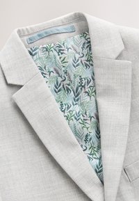 Blazer gris clair avec revers cranté dévoilant une doublure intérieure à motif de feuilles botaniques vertes et bleues ainsi qu'un design floral.