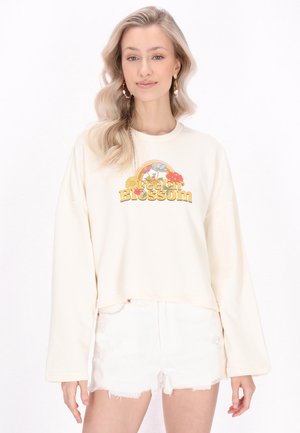 Ung kvinde med langt blondt hår bærer en cremefarvet sweatshirt med teksten "Feelin Blossom" og blomstermotiv, kombineret med hvide denimshorts.