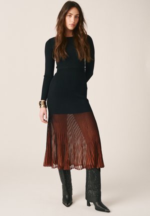 Vrouw draagt een zwarte jurk met lange mouwen en een doorschijnende geplooide rok over roestkleurige stof, zwarte laarzen met studs en een gouden polsmanchet.