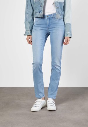 Personne portant un jean skinny bleu clair, une veste en denim, une chemise blanche et des baskets blanches avec des bandes Velcro, se tenant sur un sol gris.