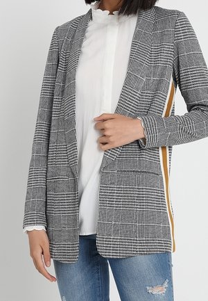 Grau-schwarzer kariert Blazer mit tailliertem Schnitt, zwei Fronttaschen und einem gelben Akzent an der Seite. Kombiniert mit einer weißen Bluse.
