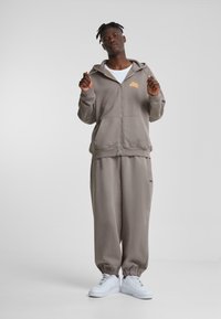 Sudadera gris con cremallera y bolsillos canguro, combinada con pantalones de chándal a juego. Zapatillas blancas completan el atuendo relajado.