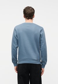 Άνδρας που φοράει γαλάζιο φούτερ με μακριά μανίκια και μαύρο παντελόνι, στέκεται με την πλάτη προς την κάμερα μπροστά σε ένα απλό λευκό φόντο.