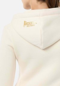 Beiger Hoodie mit einem goldenen "BOXEURS DES RUES"-Logo auf dem Rücken, aus einem strukturierten Stoff und einer weichen, glatten Kapuze.