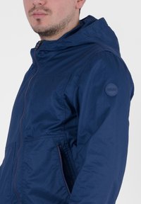 Giacca con cappuccio di colore blu navy realizzata in tessuto leggero, dotata di zip frontale, tasche laterali e dettaglio con logo rotondo sulla manica.