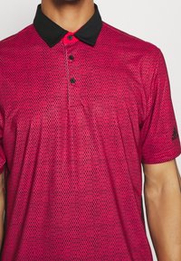 Červená a černá polo košile s krátkým rukávem a texturovaným vzorem, tři černé knoflíky a černý límec. Obsahuje logo Adidas na rukávu.